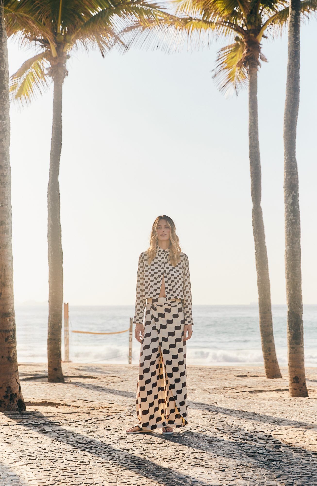 A woman in a black and white check set walking under palm trees on the beach