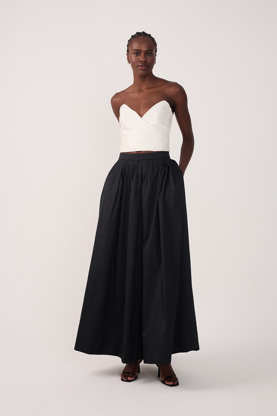 Woman wearing a white strapless sweetheart top and black pleated maxi skirt with hands in pockets