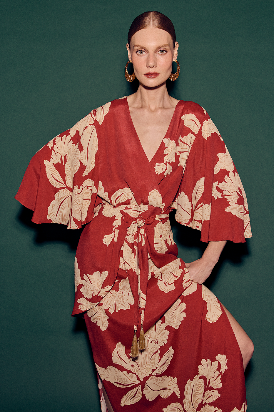 Woman in red and beige floral print kimono-sleeve wrap dress with tassel belt on dark green backdrop