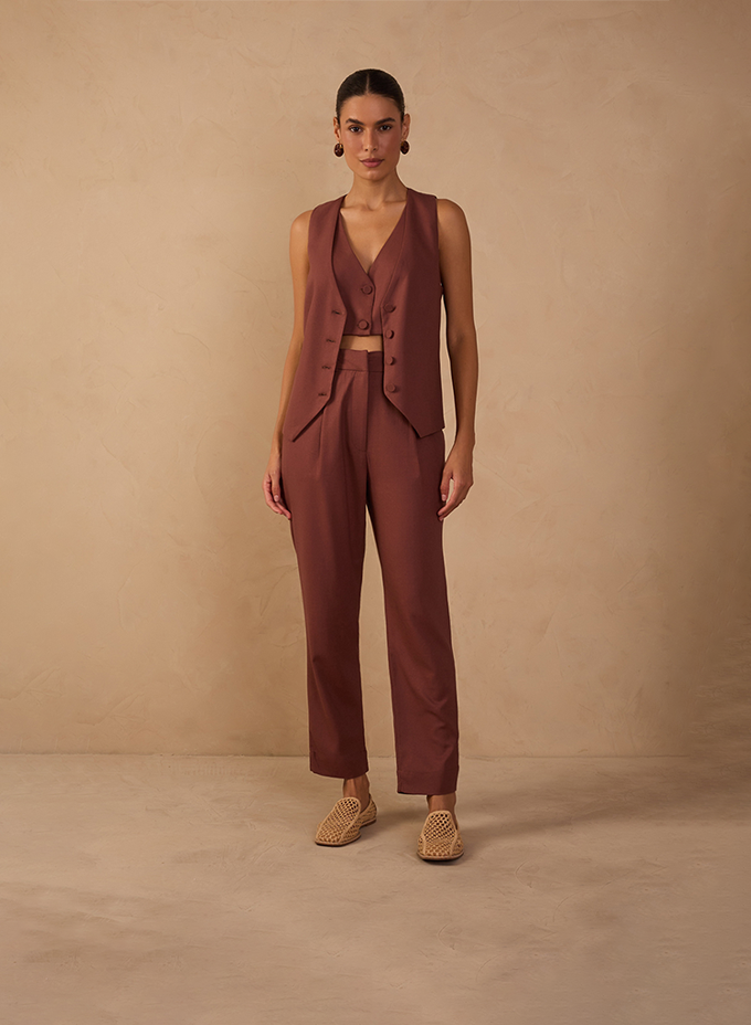 A woman stands against a warm beige textured backdrop wearing a tailored sleeveless vest and matching high-waisted trousers in a rich brown tone. The look features a cropped top underneath, clean lines, and woven flat shoes, creating a refined yet relaxed modern silhouette.