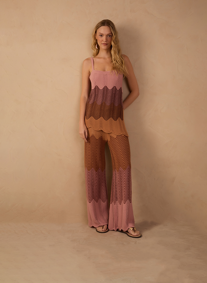 A woman stands against a warm beige textured backdrop wearing a sleeveless knit top and matching wide-leg pants. The set features a wave-pattern design in layered tones of blush pink, caramel, and brown, styled with flat sandals for a relaxed, resort-ready look.