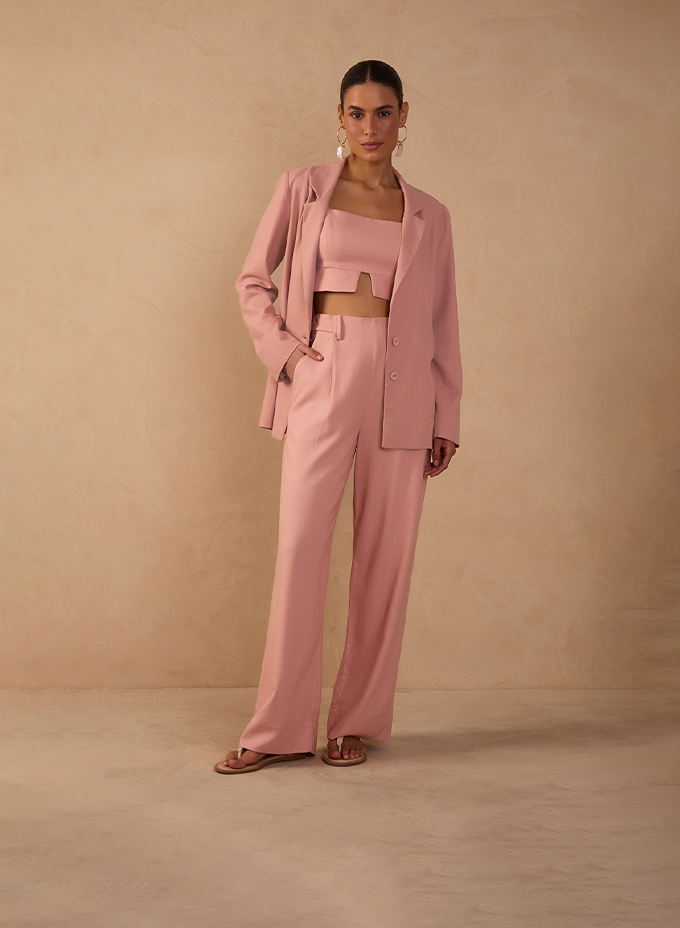 A woman stands against a warm beige textured backdrop wearing a soft pink tailored suit. The look includes a relaxed blazer, cropped top, and high-waisted wide-leg trousers, styled with flat sandals and statement earrings for a modern, feminine silhouette.