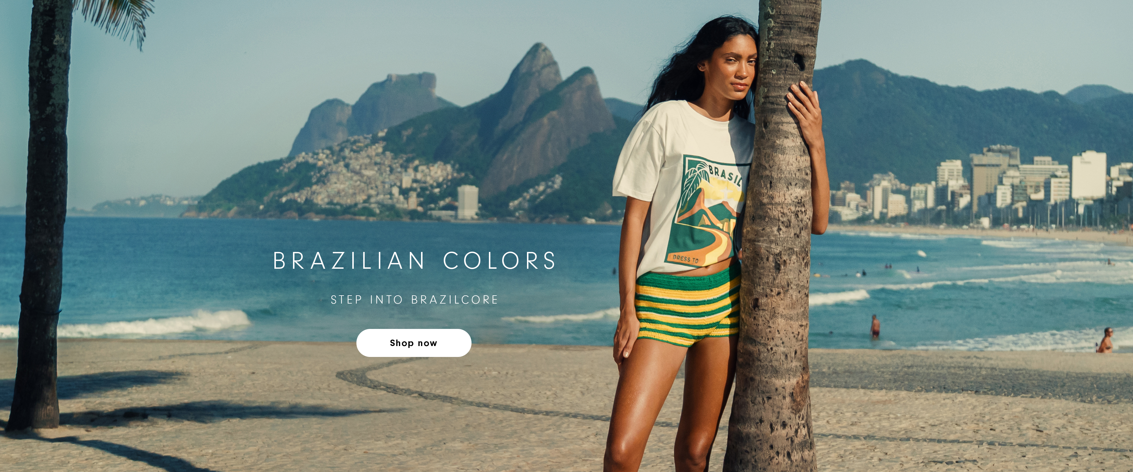 Brazilian Colors desktop banner - Step into Brazilcore - woman in white t-shirt and green and yellow striped shorts leaning against a palm tree on Ipanema beach with Two Brothers mountain in the background, Shop Now button