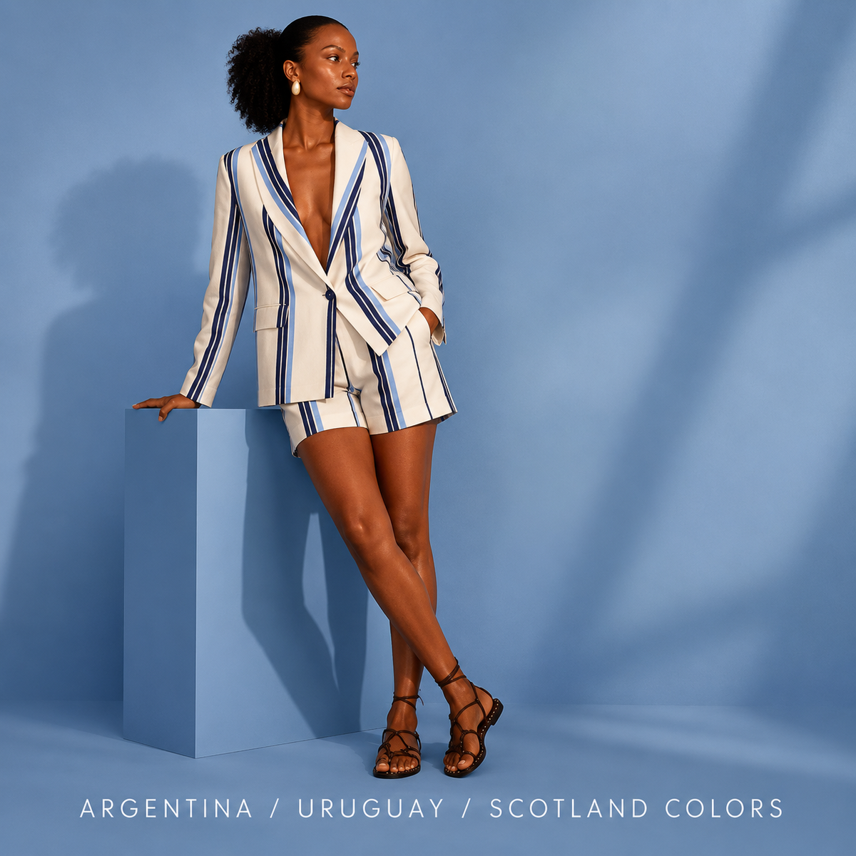 Woman wearing a white and blue striped blazer and matching shorts, leaning on a blue cube against a blue background, with the text "Argentina / Uruguay / Scotland Colors"