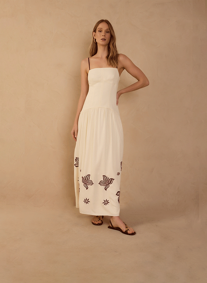 New dresses category banner: woman in an off-white strappy maxi dress with dark floral embroidery and flat sandals against a beige backdrop