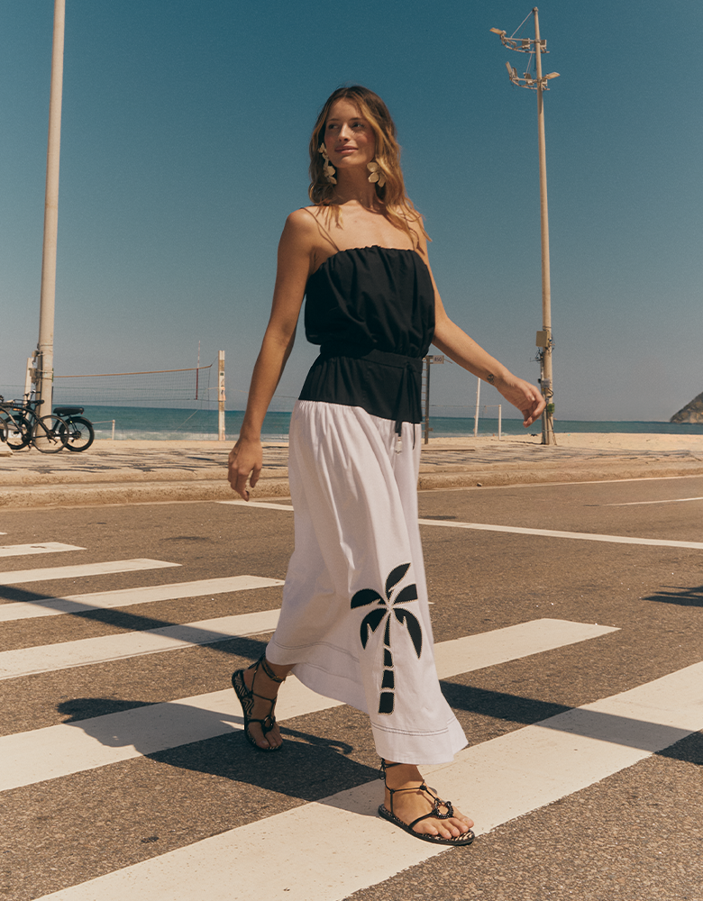 A woman wearing a black strapless top and a white maxi skirt with a black palm tree print walks across a crosswalk near a beach.