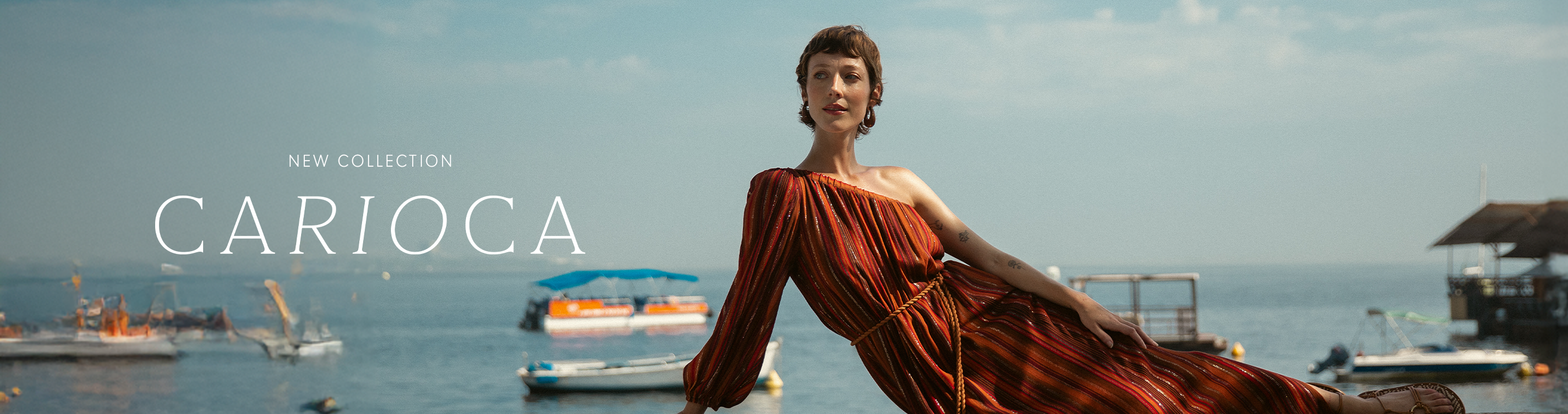Horizontal banner advertising the 'New Collection Carioca.' A model in a flowing, one-shoulder reddish-orange dress is lounging on a pier or boat deck with a calm, sunny ocean and cityscape in the background.