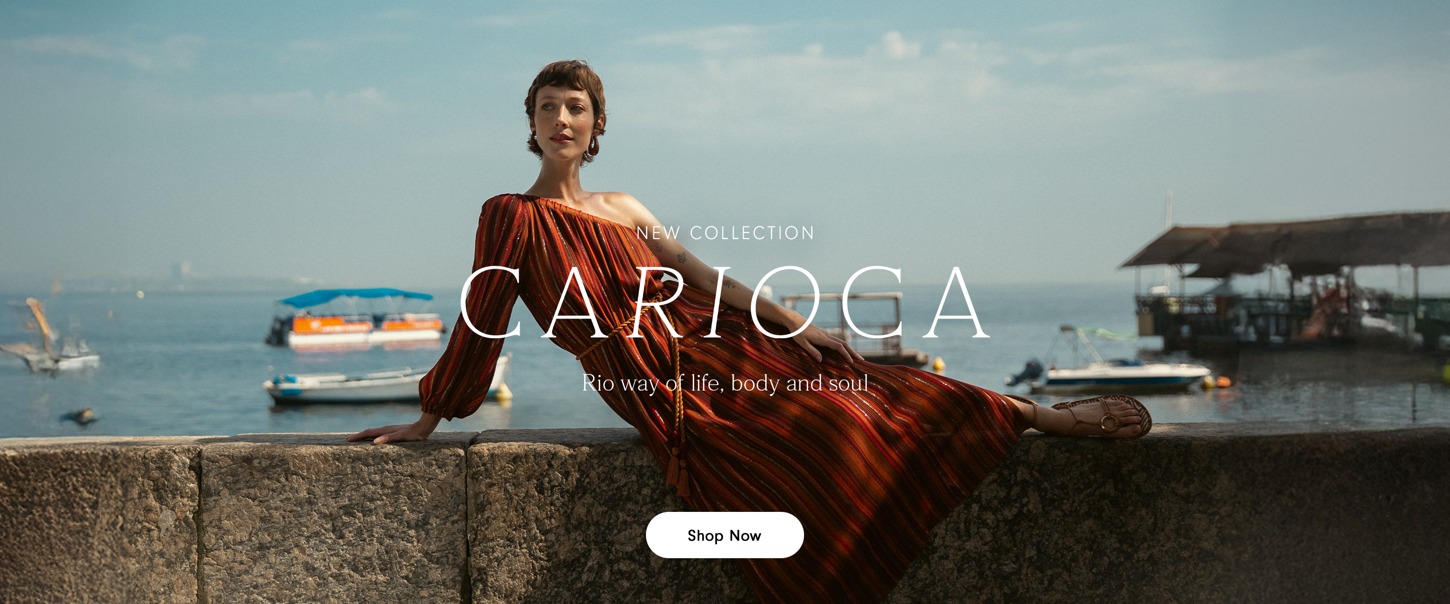 Wide banner for the 'New Collection Carioca: way of life, body and soul.' Features a woman in a flowing, reddish-orange dress relaxing on a stone wall overlooking the water. Includes a 'Shop Now' button.