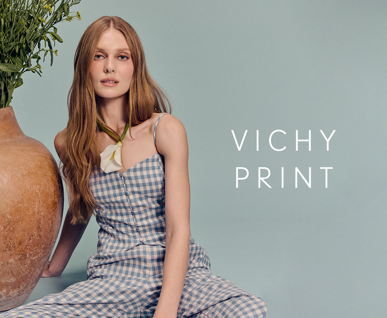 Dress To Vichy Print collection banner featuring a model in a blue and white gingham dress seated next to a ceramic vase on a teal background