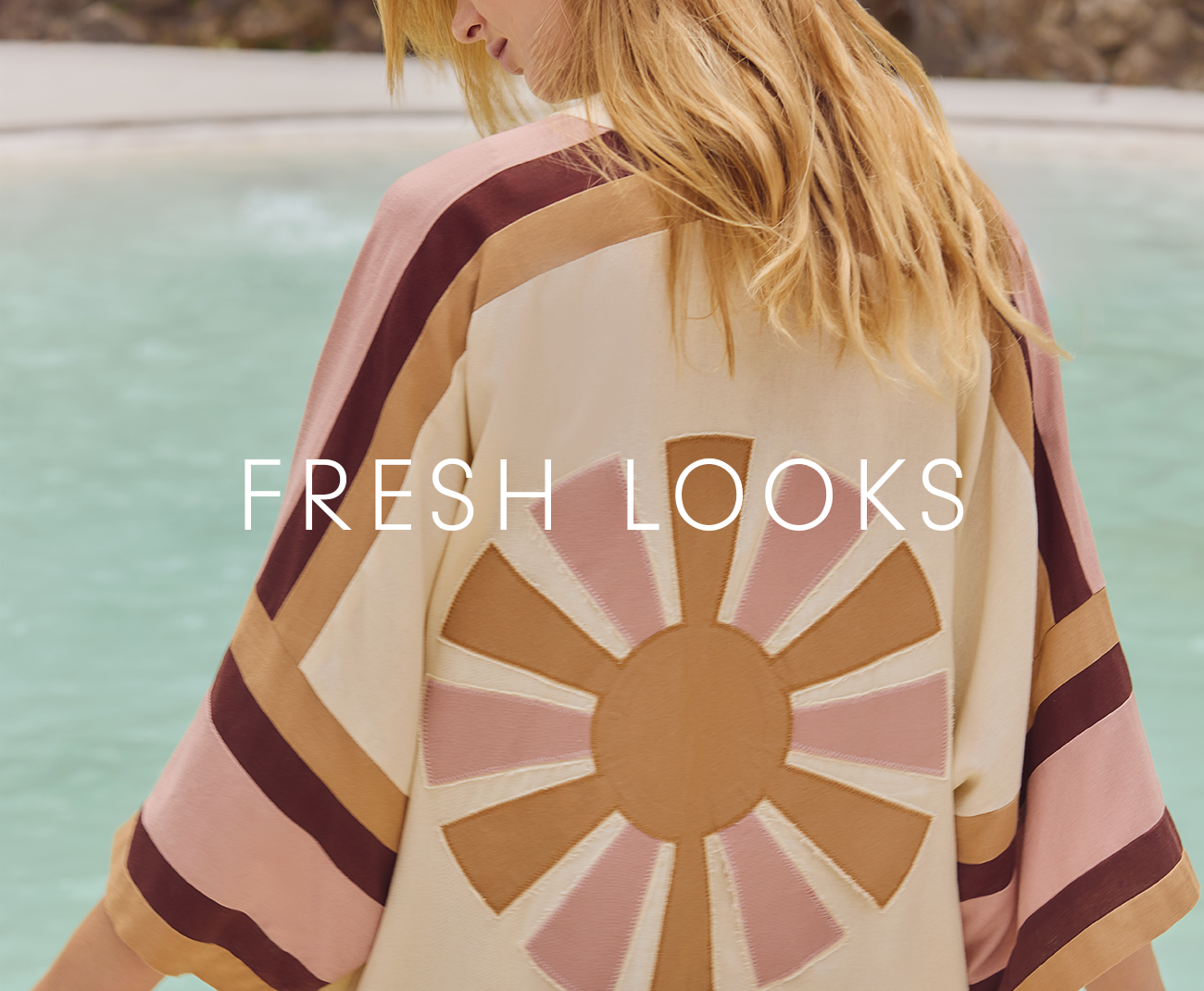 Model seen from behind wearing a cream poncho with a sunburst design in brown and pink tones near the ocean, Fresh Looks category banner for Dress To
