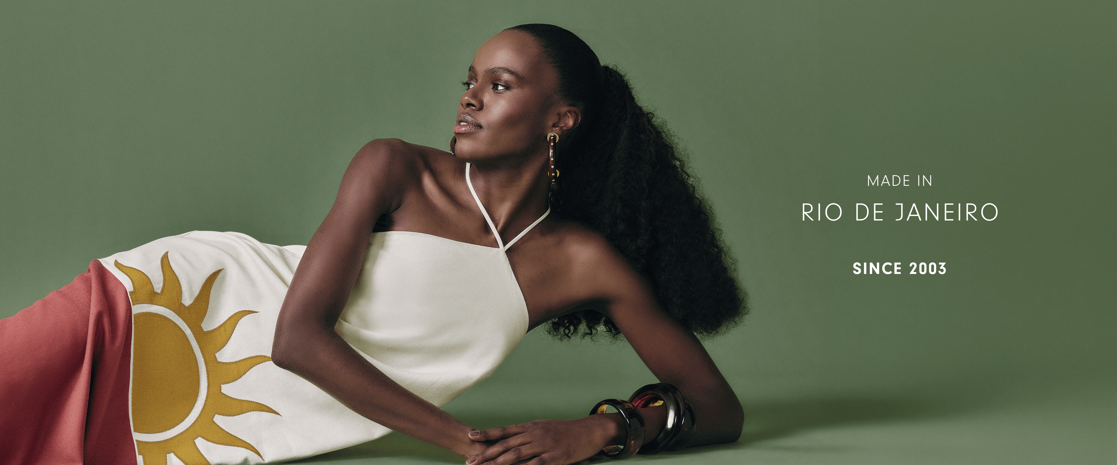 "Made in Rio de Janeiro since 2003" desktop banner: model lying on a green backdrop wearing a cream halter dress with a yellow sun print