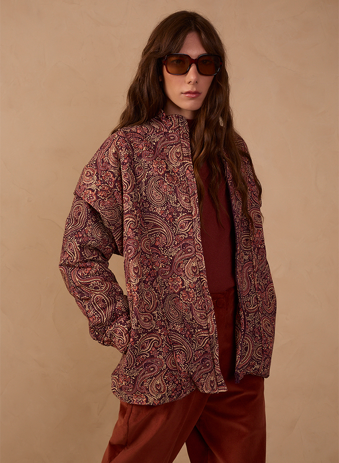 Woman wearing a burgundy paisley print quilted jacket over a rust-red top and matching wide-leg trousers, styled with tinted brown sunglasses against a warm beige backdrop.