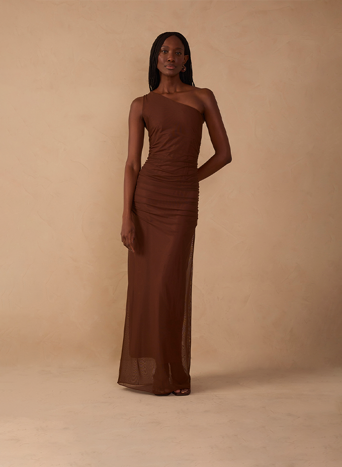 Modelo usando vestido longo marrom de um ombro só, com tecido drapeado, em fundo bege.