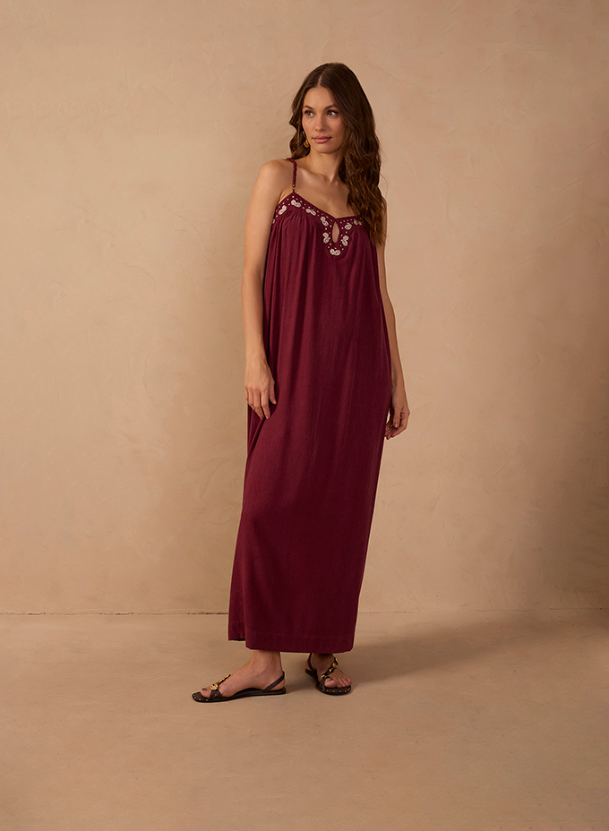 Woman wearing a burgundy sleeveless maxi slip dress with thin straps and a floral embroidered neckline, paired with black strappy flat sandals against a warm beige backdrop. Long Dresses.