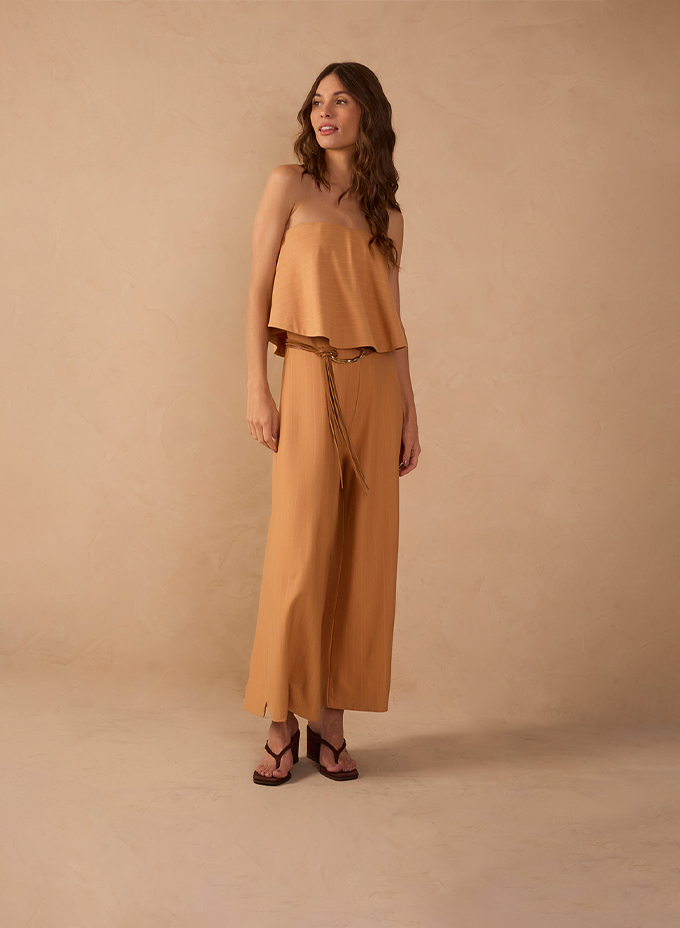 Modelo usando conjunto caramelo de blusa cropped tomara que caia e calça pantalona, em fundo bege.