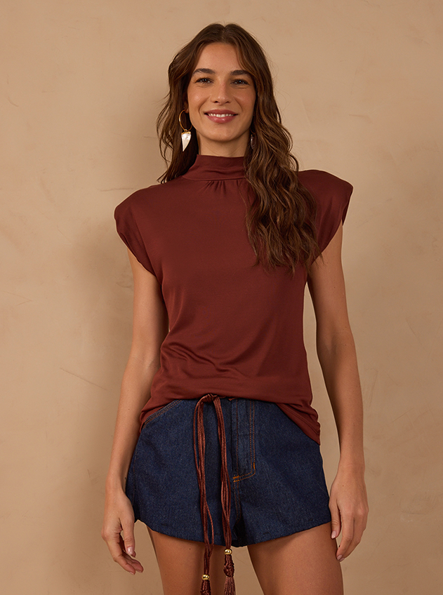 Smiling woman in a rust brown high-neck top with shoulder pads and dark denim shorts with a tie belt against a beige wall