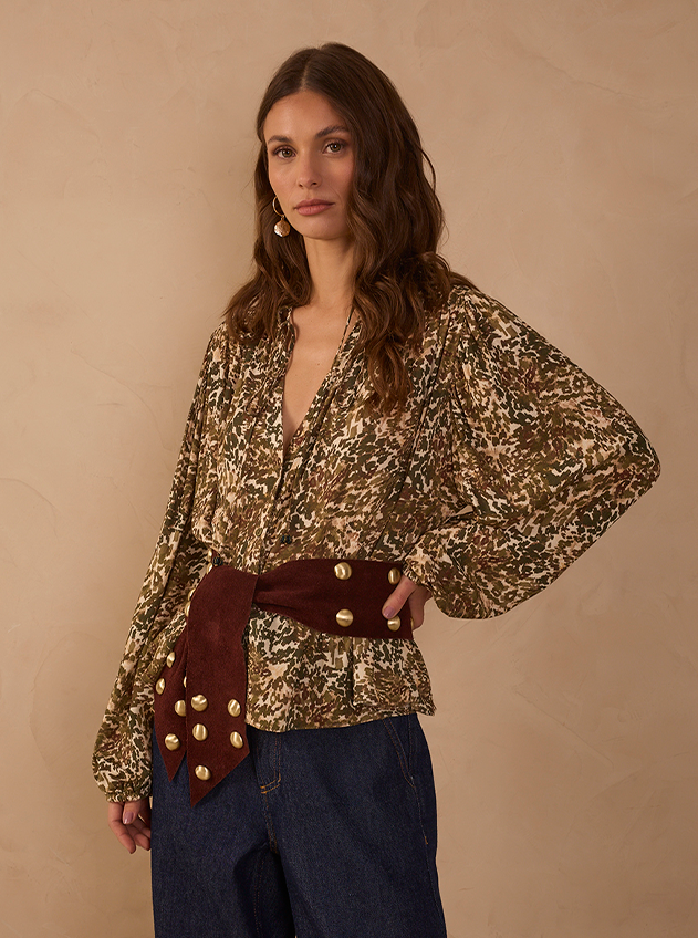 Model wearing an olive and cream abstract-print blouse with balloon sleeves, a burgundy suede sash belt with gold studs and dark wide-leg jeans, against a beige backdrop