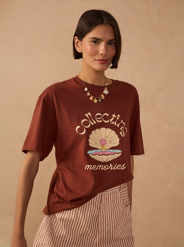 Woman wearing a brown Collecting Memories graphic t-shirt with a seashell print, paired with striped beige shorts and a layered gold necklace