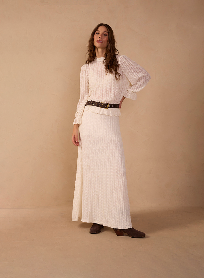 Woman wearing an off-white textured knit set with a high-neck ruffled blouse and matching maxi skirt, cinched with a brown studded belt and paired with dark brown ankle boots against a warm beige backdrop. Matching sets.