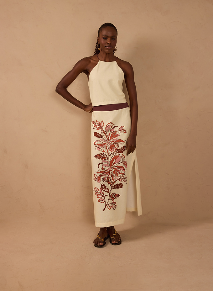 Woman in a cream halter top and long cream skirt with a large red floral embroidery and brown sandals