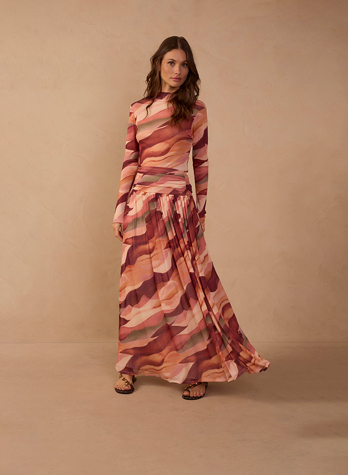 Woman wearing a long-sleeve top and matching pleated maxi skirt in a coral, pink and burgundy wavy abstract print, paired with strappy flat sandals against a warm beige backdrop.