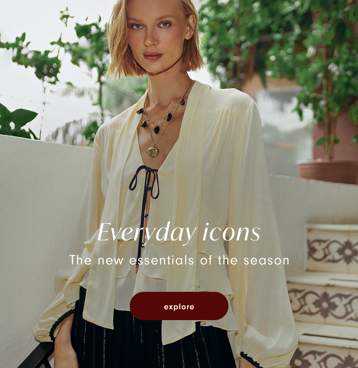 Everyday Icons banner with a model in a cream tie-front blouse, layered pendant necklaces and black pleated skirt by patterned tile steps