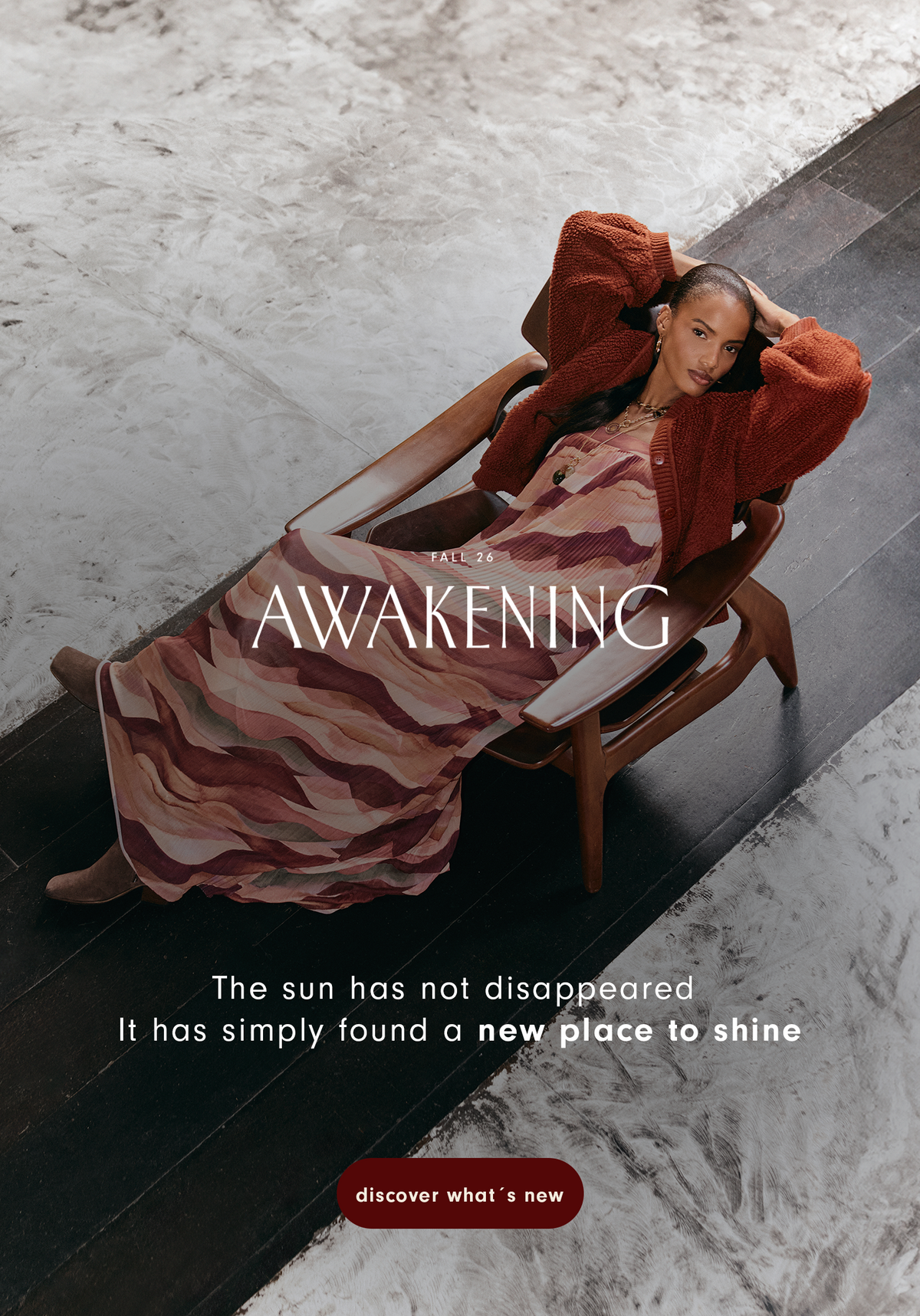 Fall 26 Awakening banner with a model in a rust cardigan and wave print maxi dress reclining in a wooden armchair on a marble floor