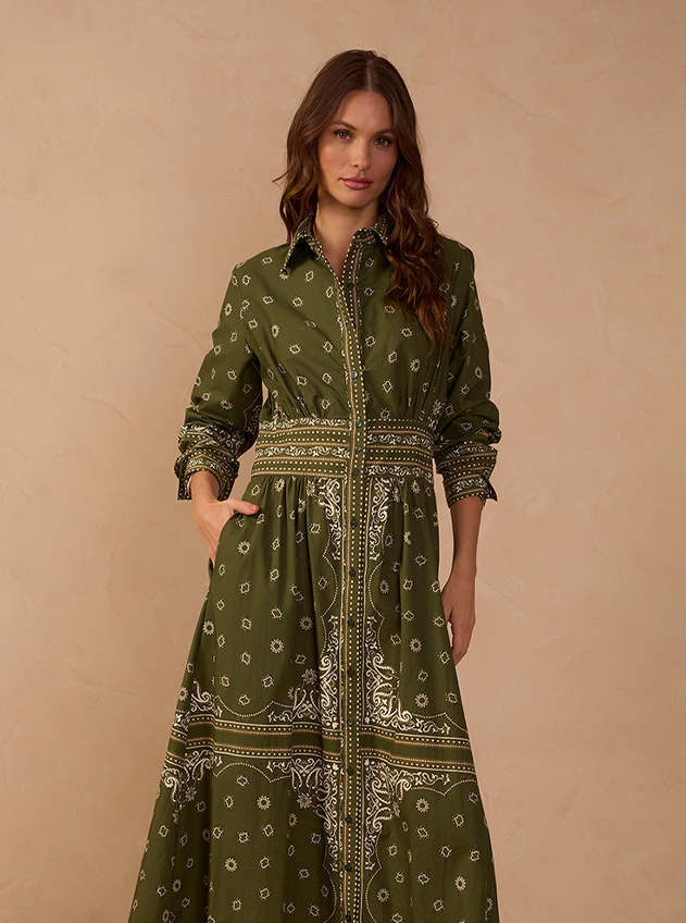 Woman wearing an olive green long-sleeve button-front shirt dress with a cream bandana paisley print, fitted waist band and flowing midi skirt against a warm beige backdrop.