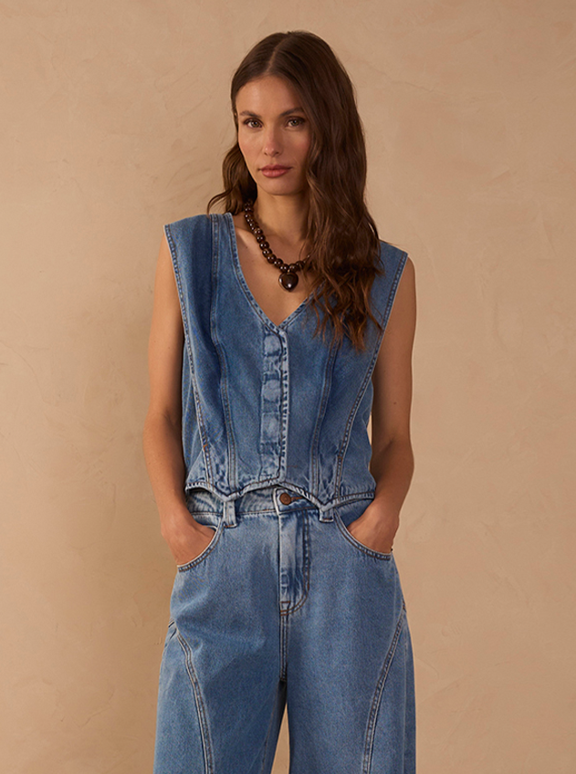 Woman wearing a sleeveless denim vest with a beaded necklace and wide-leg jeans against a beige wall