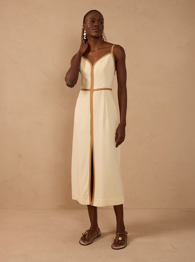 Model wearing a cream sleeveless midi slip dress with camel trim and a front slit, paired with brown leather sandals, against a beige backdrop