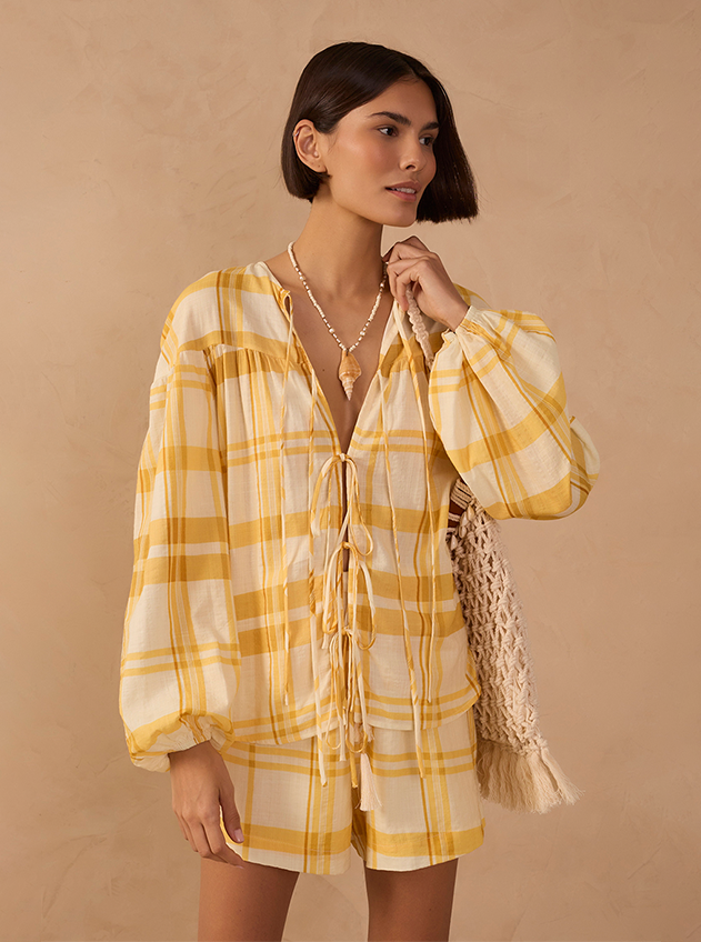 Woman wearing a yellow and white plaid blouse and shorts set with a beaded necklace, holding a crochet handbag.