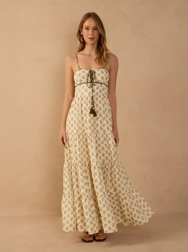Model wearing a cream tiered maxi dress with a small olive block print, thin straps and a tasselled drawstring tie, paired with black studded sandals, against a beige backdrop