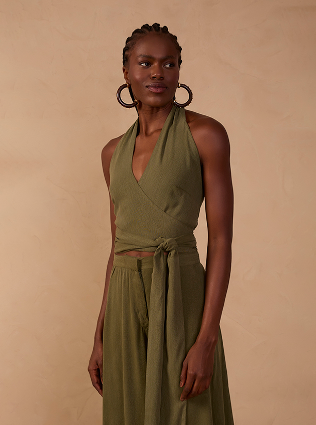 Woman wearing an olive green halter-neck wrap crop top with a tie waist and matching wide-leg trousers, styled with large gold hoop earrings against a warm beige backdrop.
