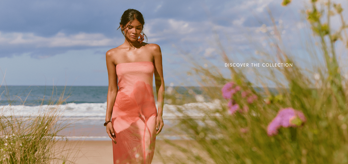 Wide lookbook banner with the text Discover the Collection, showing a model in a coral slip dress walking through beach grass by the sea