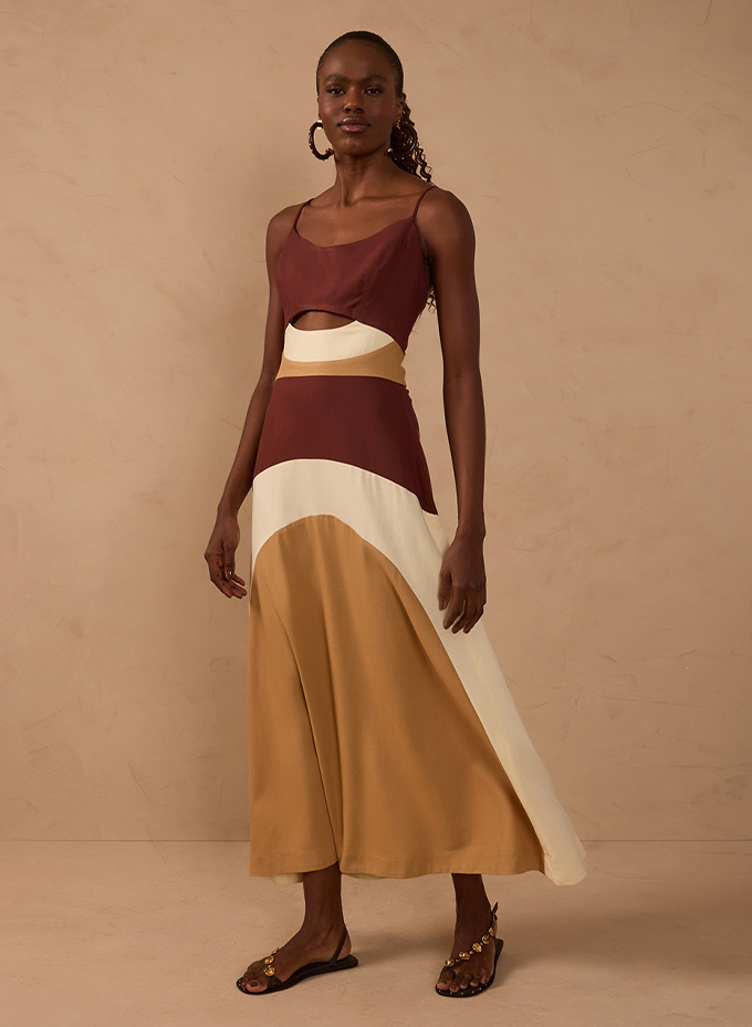 Model wearing a colour-blocked slip midi dress in burgundy, cream and camel with a waist cutout, paired with gold-embellished flat sandals, against a beige backdrop