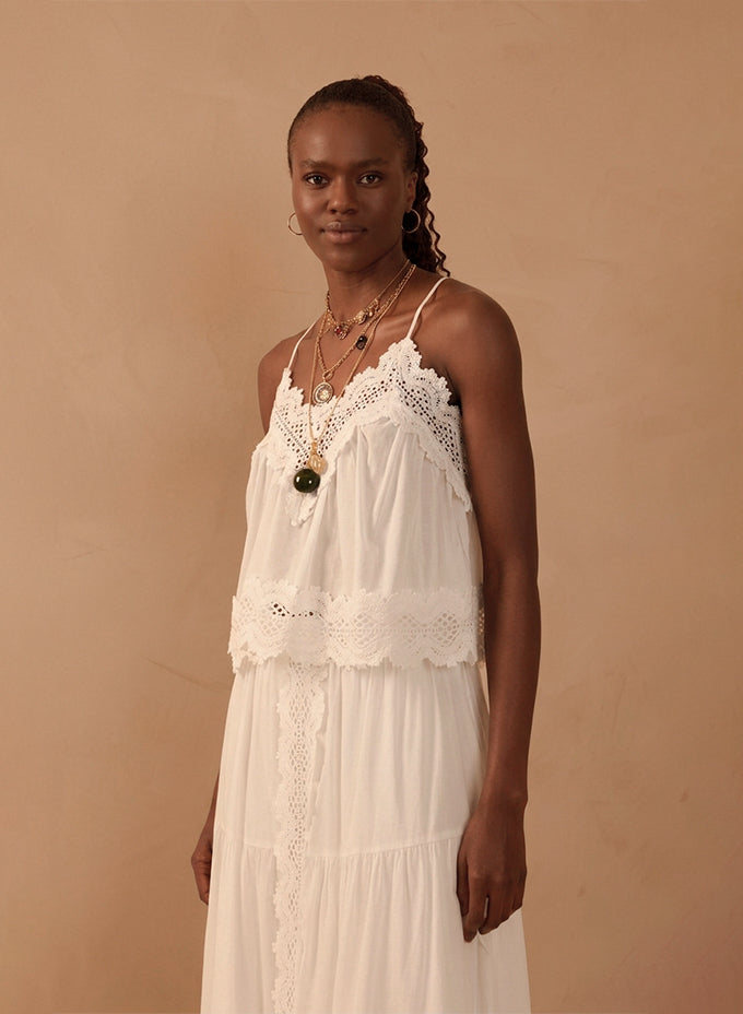 Model wearing a white lace-trimmed strappy camisole top and matching tiered maxi skirt with layered gold necklaces, against a beige backdrop