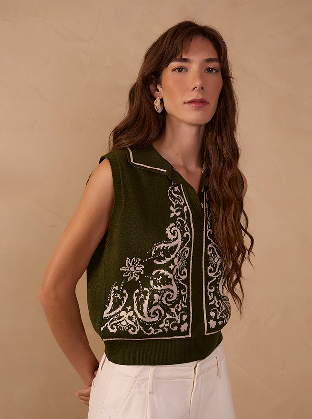 Woman wearing a dark olive green sleeveless knit polo vest with a cream paisley jacquard pattern and contrast collar trim, paired with white high-waisted trousers against a warm beige backdrop.