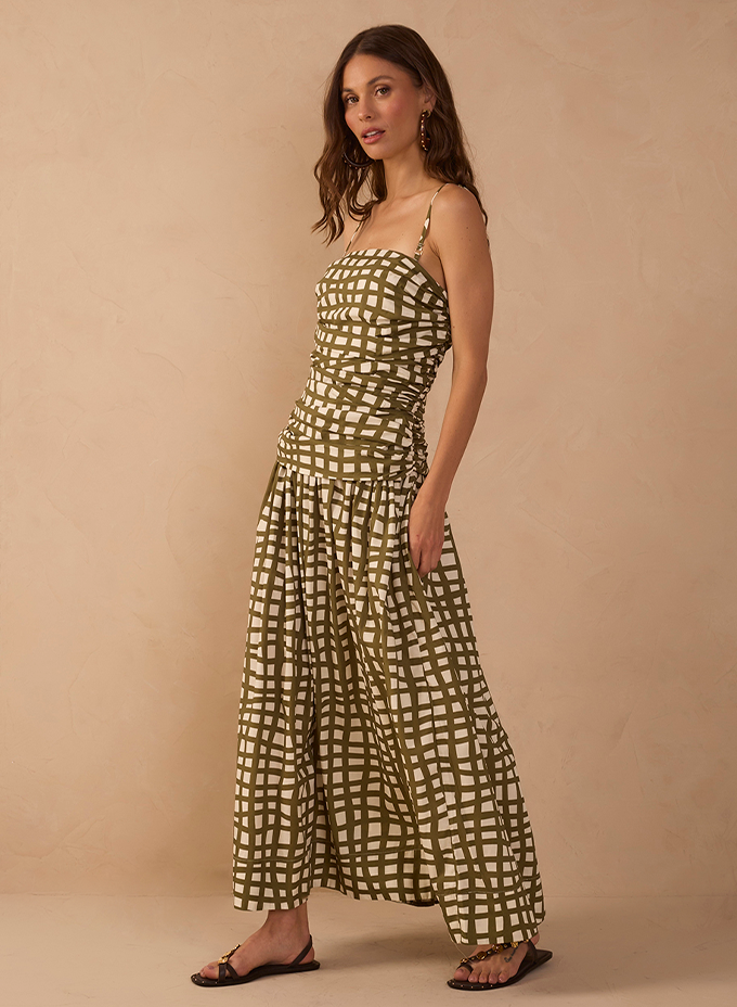 Model wearing an olive green and cream grid-print maxi dress with thin straps and a ruched bodice, paired with flat sandals, against a beige backdrop