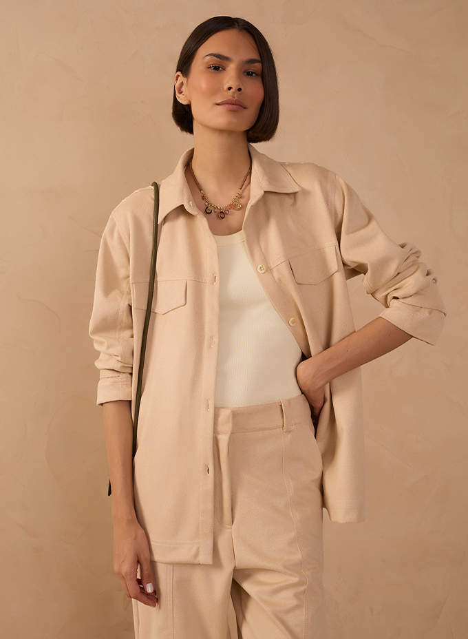 Model wearing a beige linen overshirt over a white tank top with matching wide-leg trousers and a layered gold necklace, against a beige backdrop